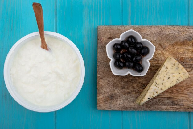 Vista superior de aceitunas negras con queso y yogur en un plato con una cuchara de madera sobre un fondo turquesa