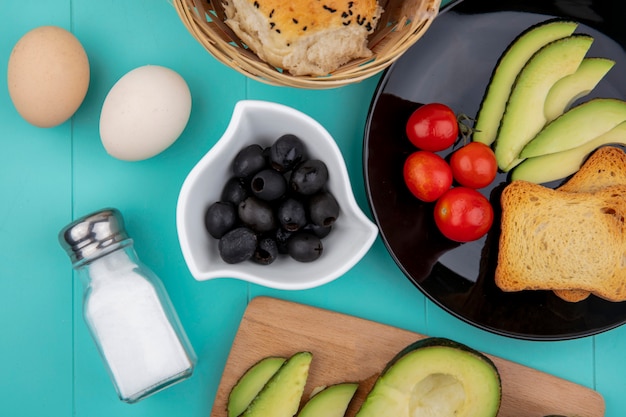 Vista superior de aceitunas negras en un tazón blanco con verduras como tomates rebanada de aguacate en un plato negro y un cubo de panes en azul