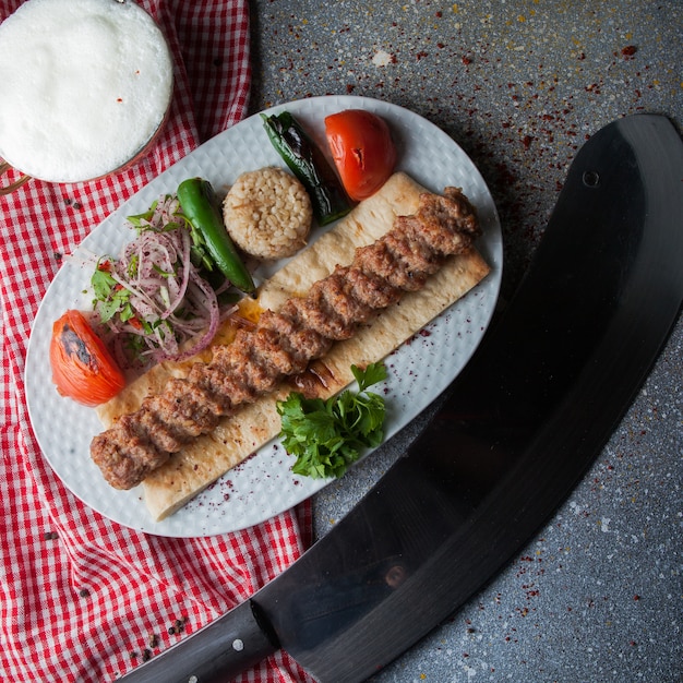 Vista superior adana kebab con cuchillo y verduras fritas y cebolla picada y ayran en plato blanco