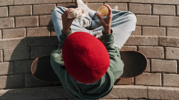 Vista superior del adolescente con patineta comiendo un sándwich y bebiendo jugo