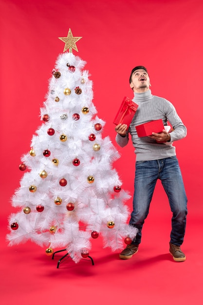 Vista superior del adulto guapo feliz en una blusa gris de pie cerca del árbol de Navidad blanco decorado