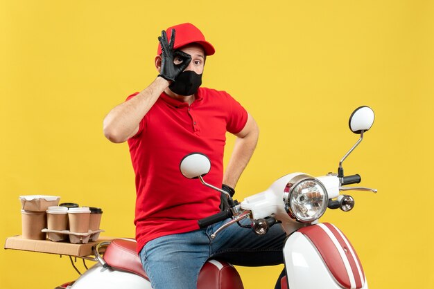 Vista superior del adulto joven con blusa roja y guantes de sombrero en máscara médica entregando orden sentado en scooter haciendo gesto de anteojos