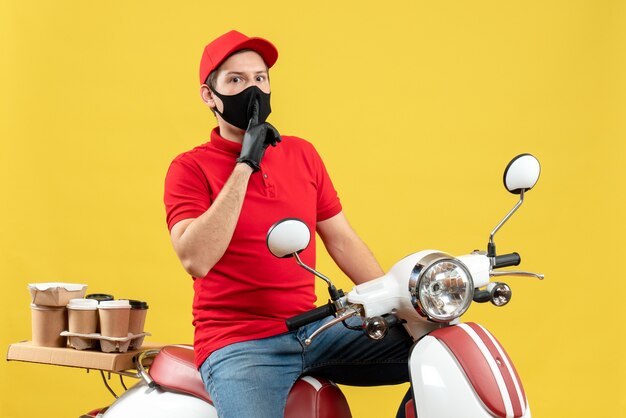 Vista superior del adulto joven con blusa roja y guantes de sombrero en máscara médica entregando orden sentado en scooter haciendo gesto de silencio sobre fondo amarillo