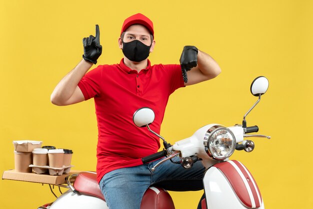 Vista superior del adulto joven con blusa roja y guantes de sombrero en máscara médica entregando orden sentado en scooter con los pulgares hacia arriba y hacia abajo sobre fondo amarillo