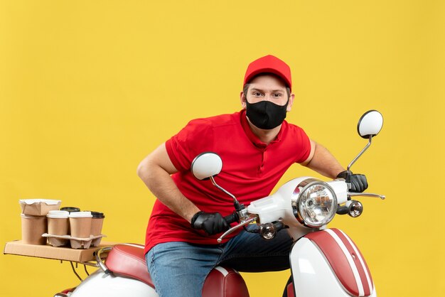 Vista superior del adulto joven confiado con blusa roja y guantes de sombrero en máscara médica entregando orden sentado en scooter sobre fondo amarillo