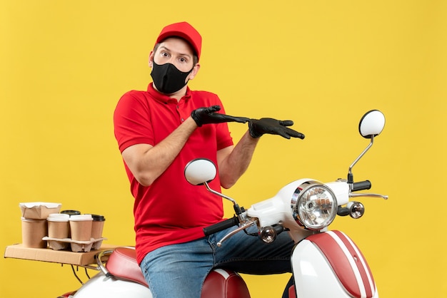 Vista superior del adulto joven emocional con blusa roja y guantes de sombrero en máscara médica entregando orden sentado en scooter apuntando algo en el lado izquierdo