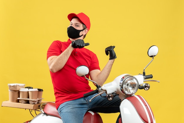 Vista superior del adulto joven emocional con blusa roja y guantes de sombrero en máscara médica entregando orden sentado en scooter apuntando hacia atrás