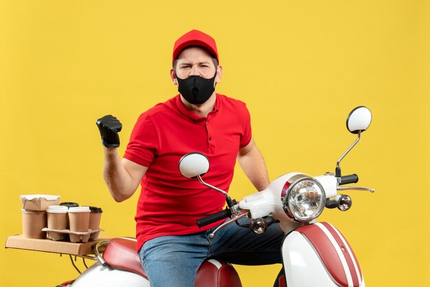 Vista superior del adulto joven emocional con blusa roja y guantes de sombrero en máscara médica entregando orden sentado en scooter apuntando hacia atrás