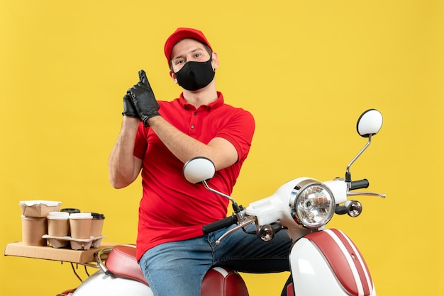 Vista superior del adulto joven emocional con blusa roja y guantes de sombrero en máscara médica entregando orden sentado en scooter haciendo gesto de pistola