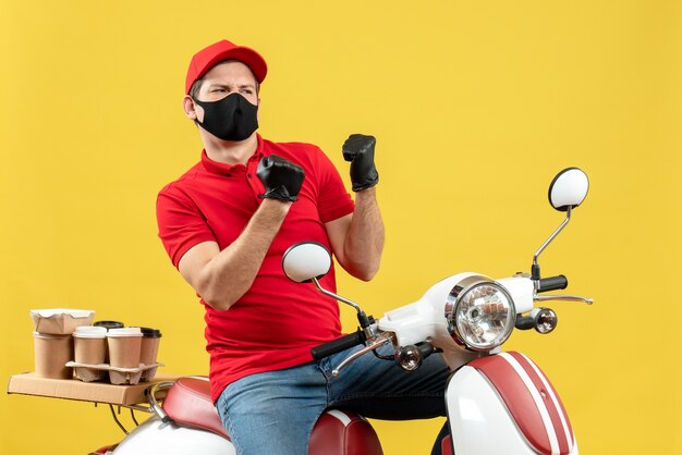 Vista superior del adulto joven emocional con blusa roja y guantes de sombrero en máscara médica entregando orden sentado en scooter sintiéndose enojado