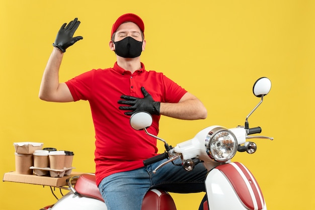 Vista superior del adulto joven emocional con blusa roja y guantes de sombrero en máscara médica entregando orden sentado en scooter sintiéndose esperanzado