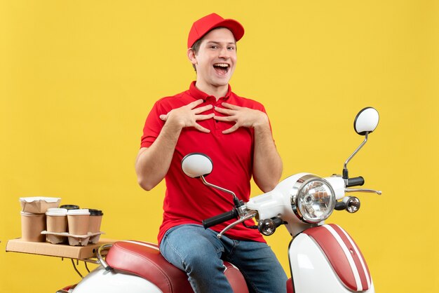 Vista superior del adulto joven inseguro inseguro sonriente que lleva la blusa roja y el sombrero que entrega el orden sentado en scooter apuntando a sí mismo en la pared amarilla