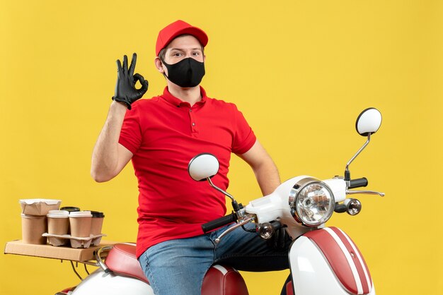 Vista superior del adulto joven orgulloso y seguro con blusa roja y guantes de sombrero en máscara médica entregando orden sentado en scooter haciendo gesto perfecto sobre fondo amarillo