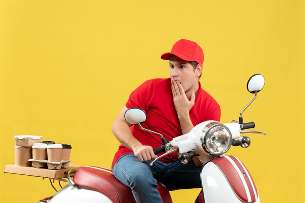 Vista superior del adulto joven pensativo con blusa roja y sombrero entregando orden en la pared amarilla