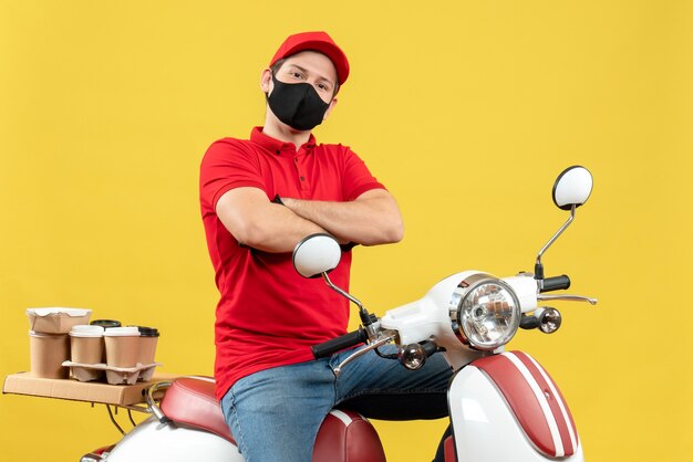 Vista superior del adulto joven reflexivo con blusa roja y guantes de sombrero en máscara médica entregando orden sentado en scooter en la pared amarilla