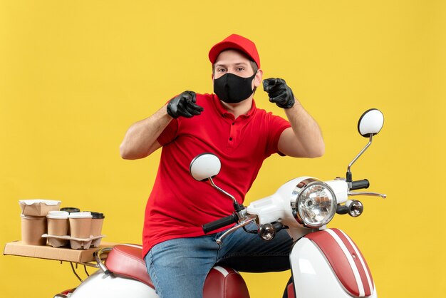 Vista superior del adulto joven sonriente con blusa roja y guantes de sombrero en máscara médica entregando orden sentado en scooter apuntando hacia adelante en la pared amarilla