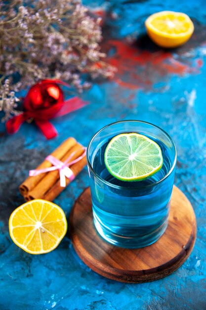 Vista superior de un agua sin gas en un vaso sobre una tabla de cortar de madera y limones, canela, limas, cono de coníferas, accesorio de decoración sobre fondo azul.