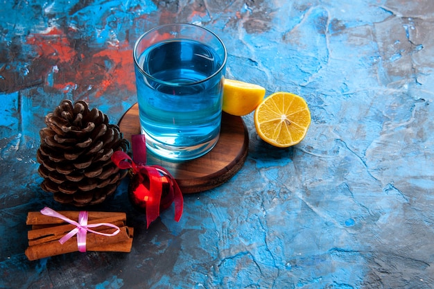 Vista superior de un agua sin gas en un vaso sobre una tabla de cortar de madera y limones, canela, limas, cono de coníferas en el lado derecho sobre fondo azul.