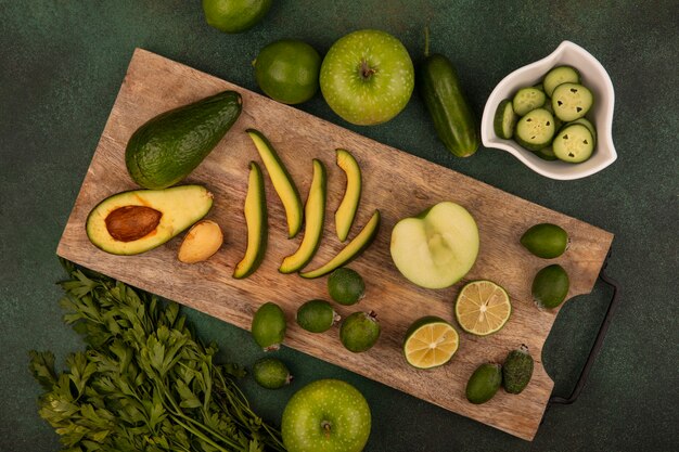 Vista superior de aguacate saludable con rodajas en una tabla de cocina de madera con feijoas medias limas con rodajas de pepino picado en un recipiente con limones manzanas verdes y perejil aislado sobre un fondo verde