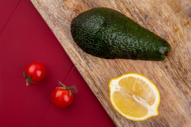 Vista superior de aguacate verde y fresco sobre una tabla de cocina de madera con rodaja de limón y tomates cherry en superficie roja