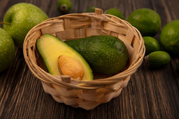 Vista superior de aguacates frescos en un balde con limas de manzanas verdes y feijoas aislado en una pared de madera