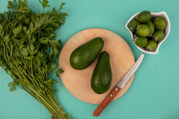 Vista superior de aguacates frescos en una tabla de cocina de madera con cuchillo con feijoas en un recipiente sobre una pared azul