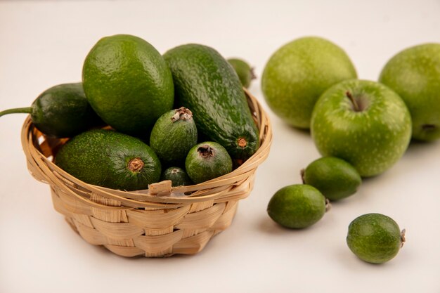 Vista superior de aguacates maduros con pepino en un balde con manzanas verdes y feijoas aislado en una pared blanca