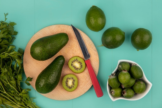 Vista superior de aguacates maduros en una tabla de cocina de madera con cuchillo con feijoas en un recipiente con limas aislado en una pared azul