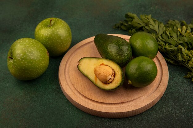 Vista superior de aguacates orgánicos en una tabla de cocina de madera con limas con manzanas verdes y perejil aislado sobre un fondo verde