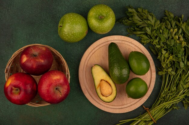 Vista superior de aguacates de piel verde sobre una tabla de cocina de madera con limas con manzanas rojas en un balde con manzanas verdes y perejil aislado sobre un fondo verde