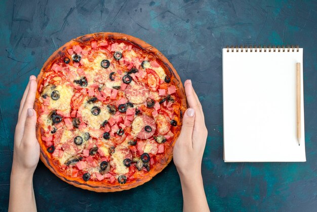 Vista superior al horno deliciosa pizza con aceitunas, salchichas y queso con bloc de notas sobre fondo azul.