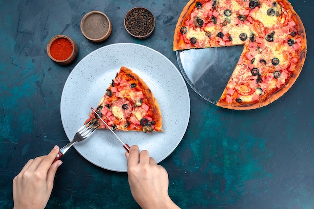 Vista superior al horno deliciosa pizza con aceitunas, salchichas y queso hembra comiéndolo en el escritorio azul.