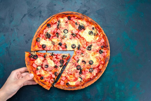 Vista superior al horno deliciosa pizza con aceitunas, salchichas y queso hembra tomando su rebanada en el escritorio azul.