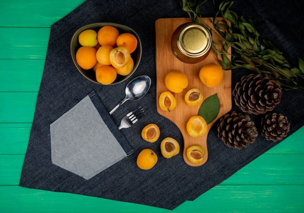 Foto gratuita vista superior de albaricoques y mermelada de durazno en tabla de cortar con tazón de albaricoques piñas hojas sobre tela de jeans con cuchara y tenedor en el bolsillo sobre fondo verde