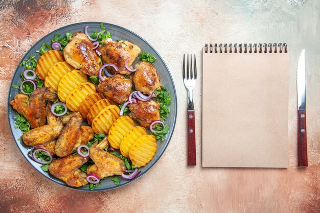 Vista superior de alitas de pollo plato de patatas pollo hierbas cebollas tenedor cuchillo cuaderno