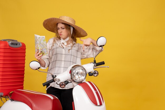 Vista superior de la ambiciosa joven con sombrero recogiendo su equipaje sentado en una motocicleta y sosteniendo el mapa haciendo un gesto de ok