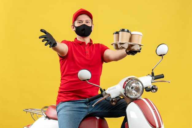 Vista superior del ambicioso repartidor sonriente con uniforme y guantes de sombrero en máscara médica sentado en scooter mostrando órdenes hablando de algo