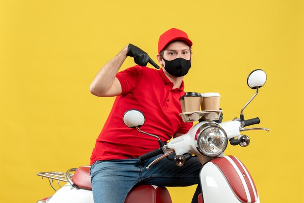 Vista superior del ambicioso repartidor con uniforme y guantes de sombrero en máscara médica sentado en scooter señalando órdenes