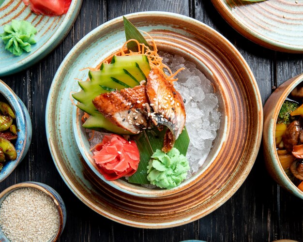 Vista superior de anguila japonesa a la parrilla con rodajas de pepinos y jengibre servidos con salsa de wasabi en hojas de bambú y cubitos de hielo en un plato sobre la mesa