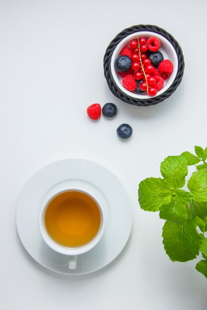 Vista superior de arándanos y frambuesas en un tazón con grosellas rojas, té de manzanilla, hojas de menta.