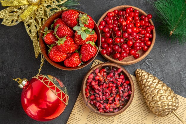 Vista superior de arándanos rojos frescos con otras frutas alrededor de juguetes navideños sobre fondo oscuro