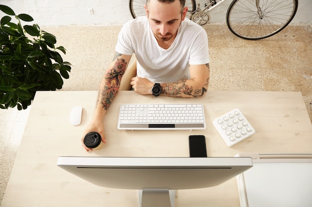 La vista superior del autónomo tatuado sostiene el café de una taza de papel para llevar mientras mira en la pantalla de su computadora con el controlador de música midi en el escritorio de su casa con un teléfono inteligente.