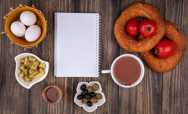 Vista superior de bagels de sésamo turco con huevos en un balde con aceitunas en un tazón y una taza de té sobre un fondo de madera con espacio de copia