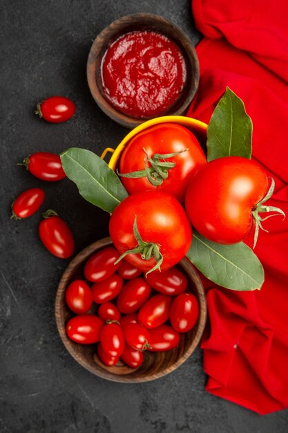 Vista superior de un balde con tomates y hojas de laurel, cuencos con tomates cherry y salsa de tomate y una toalla roja sobre suelo oscuro