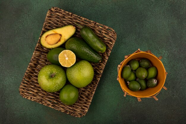 Vista superior de una bandeja de mimbre de alimentos frescos como manzanas verdes, limas, aguacate y pepino con feijoas en un balde sobre un fondo verde