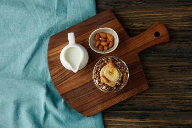 Vista superior de batido con mantequilla de maní de leche de nuez de almendras de plátano en vidrio y leche con almendras en tabla de cortar sobre tela azul sobre fondo de madera