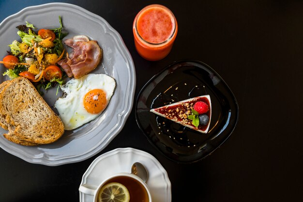 Vista superior del batido; tarta de queso; té; brindis; ensalada; tocino; Huevo frito y pan tostado en un plato gris sobre fondo negro