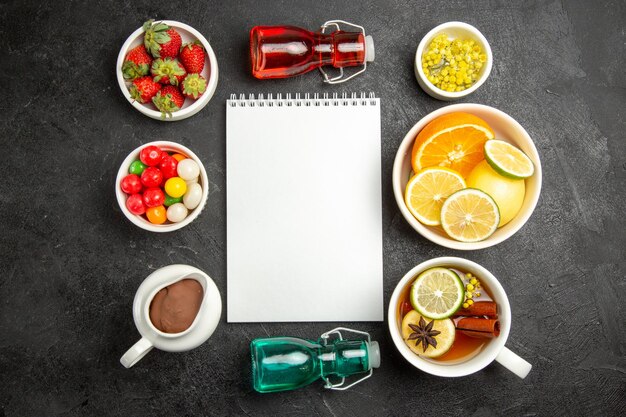 Vista superior de bayas lejanas y tazones de chocolate de crema de chocolate, dulces, hierbas, fresas y limones en rodajas junto al cuaderno blanco, la taza de té y botellas de colores