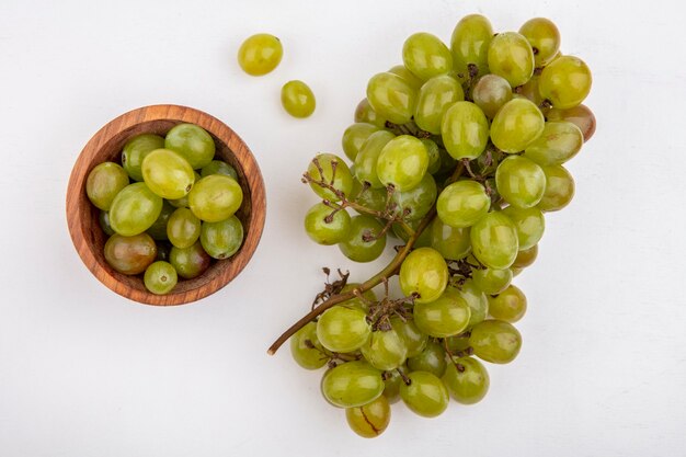 Vista superior de las bayas de uva blanca en un tazón y un racimo de uva blanca sobre fondo blanco.