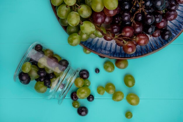 Foto gratuita vista superior de las bayas de uva que se derraman fuera del frasco de vidrio con uvas en placa sobre fondo azul.